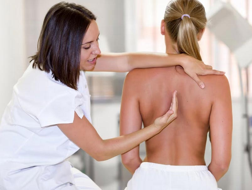 A female professional chiropractor is holding the shoulders of another female with one hand and the female chiropractor is using her other hand to explain to the other female in her care that the area she is pointing to is causing her back discomfort  between both her shoulder blades and that she will need professional chiropractor help to get posture and scoliosis care.